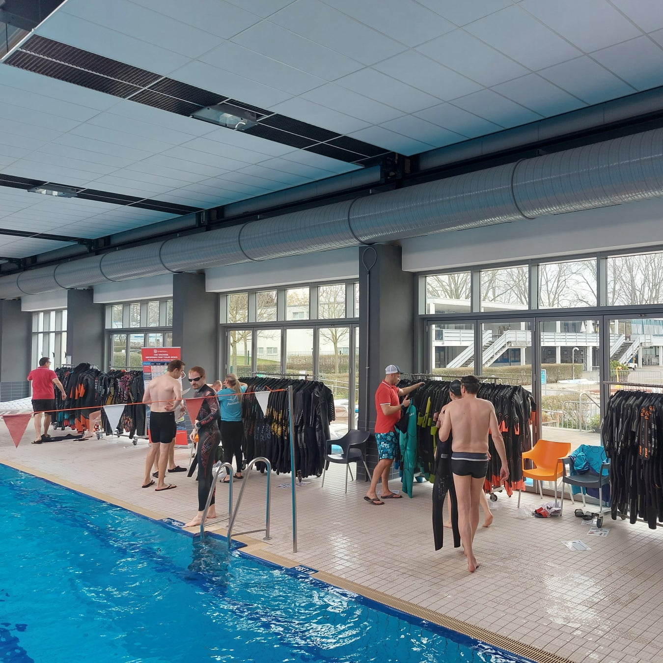Neoprentest in einer Schwimmhalle: viele Neoprenazüge hängen aufgereiht auf mehreren Ständern. Sportler ziehen die Anzüge an oder aus, gehen ins Wasser oder kommen aus dem Wasser.