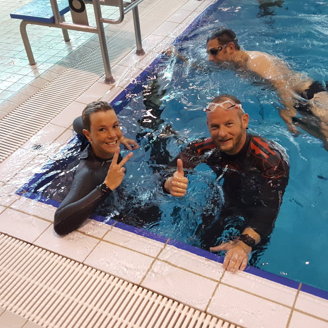 Ein Mann und eine Frau im Wasser am Beckenrand einer Schwimmbahn. Sie tragen einen Neoprenanzug, lächeln in die Kamera.