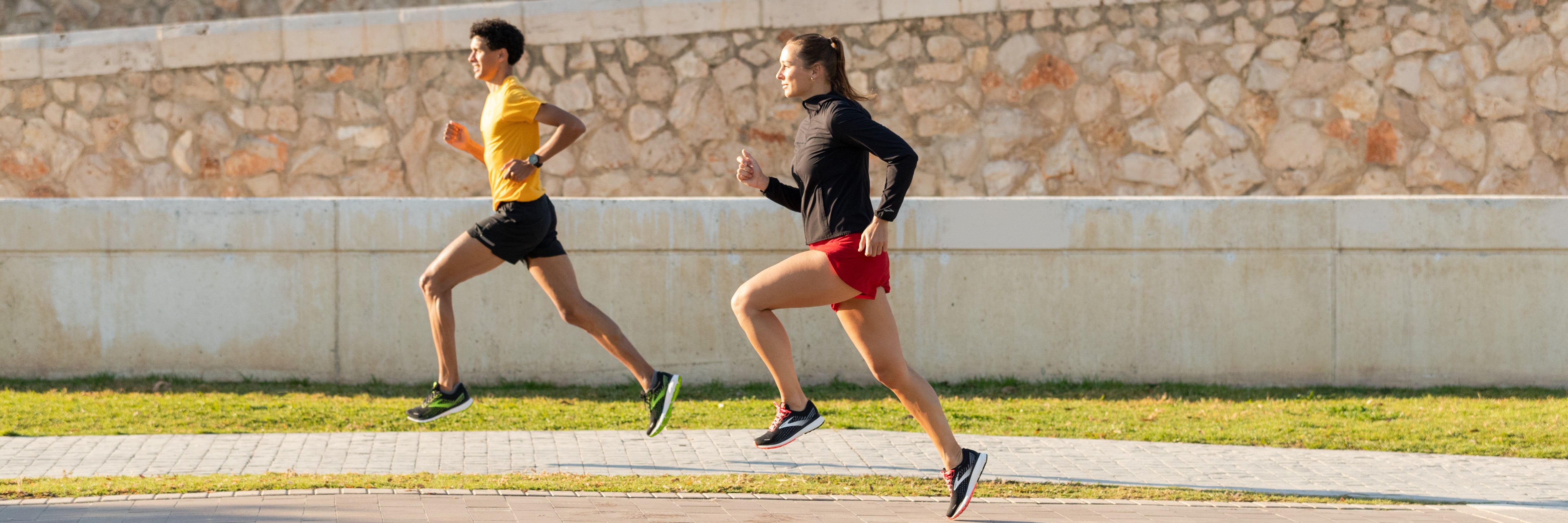 Ein Läufer und eine Läuferin von der Seite laufen auf einem sonnigen Weg an einer Mauer entlang